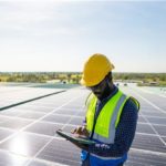 A person in a hard hat reviewing field service software on a tablet, while surrounded by solar panels